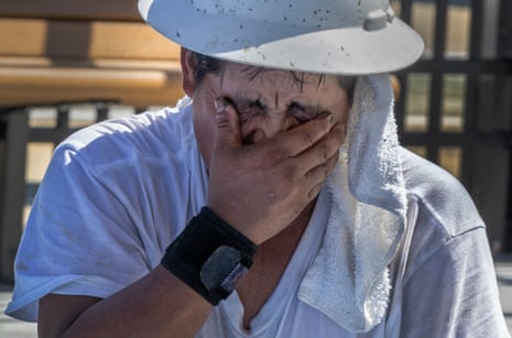 A man in a hard hat wipes sweat from his eyes with his hand, a towel around his neck
