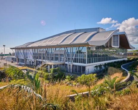 Exterior of the arrivals terminal, which has a curved roof and glass facade. Plants are growing in the foreground surrounding the building